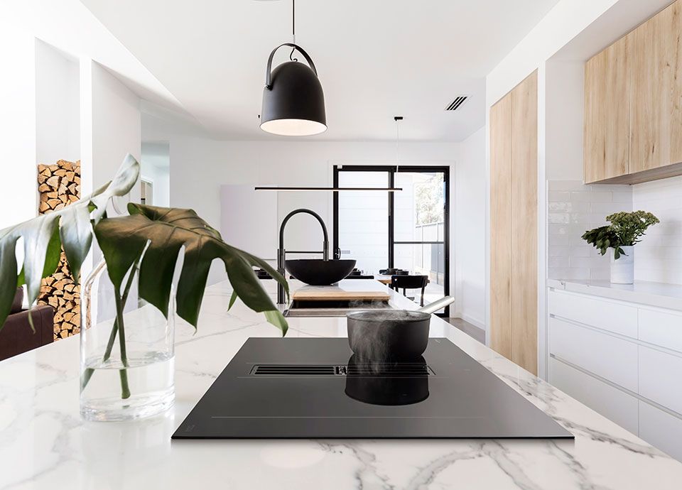 A bright, white kitchen with white units, marble worktop, and wood effect cabinetry. In the centre, is an island unit with white marble worksurface and integrated induction hob with built-in extraction