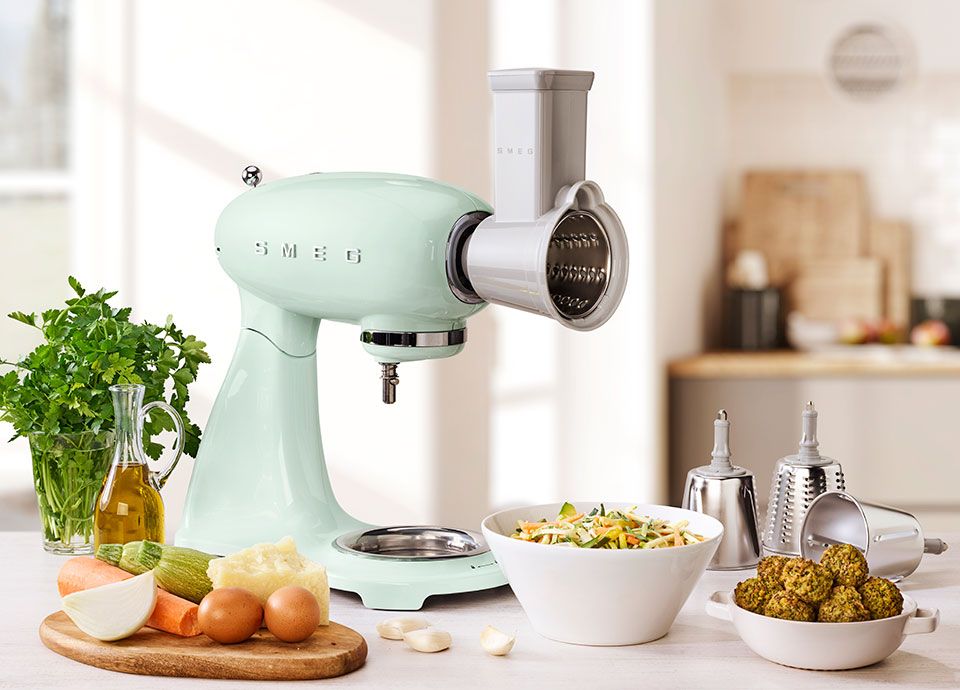 A grey smeg food mixer with a vegetable cutter accessory attached to the front, and beneath it, is a bowl of freshly cut vegetables such as carrots, peppers and cucumbers