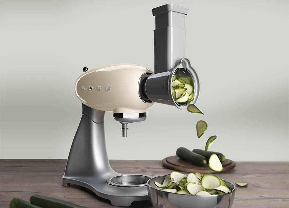 A grey smeg food mixer with a vegetable cutter accessory attached to the front, and beneath it, is a bowl of freshly cut vegetables such as carrots, peppers and cucumbers