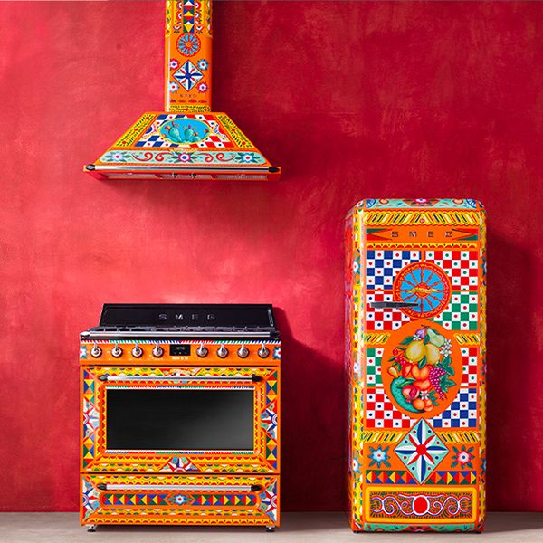 A vivid red kitchen showcasing Smeg's Divina Cucina range cooker, freestanding fridge, and chimney hood. Each in a visually stunning hand painted design, with a primarily orange base colour
