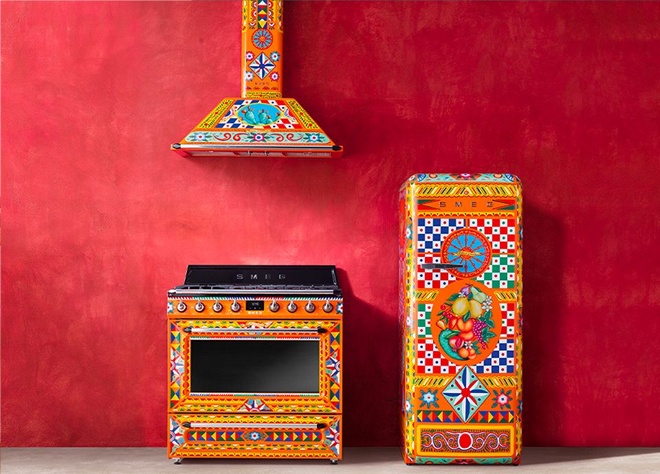 A cooker, fridge, and chimney hood from our 'Divina cucina' stand in front of a vivid red wall. The appliances feature an intricate design and are predominantly orange in colour with hints of blue and white