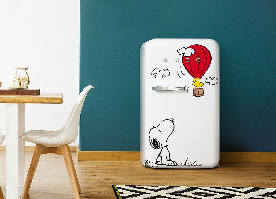 A small, white freestanding fridge featuring an illustration of Snoopy (a dog) sat in the bottom left, looking towards his friend Woodstock (a yellow bird) flying in a red hot air balloon