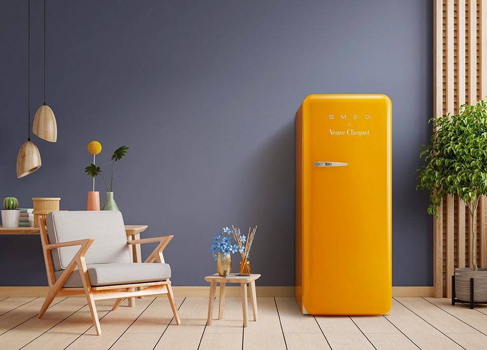 A freestanding fridge in Smeg's iconic rounded edge retro design, in Veuve Clicquot brand yellow, set against a steel blue/grey wall
