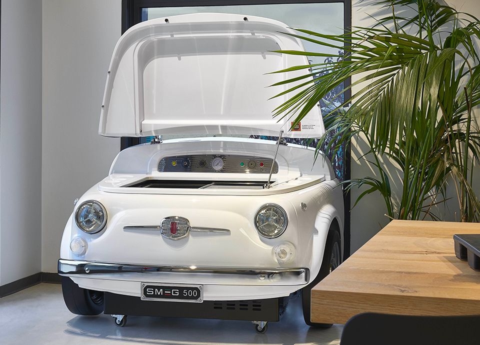 A white Smeg 500 refrigerator. This stunning appliance resembles the front end of a Fiat 500 car. The bonnet is open, revealing the top opening cooler within. Perfect for the office or adding a wow factor to any room