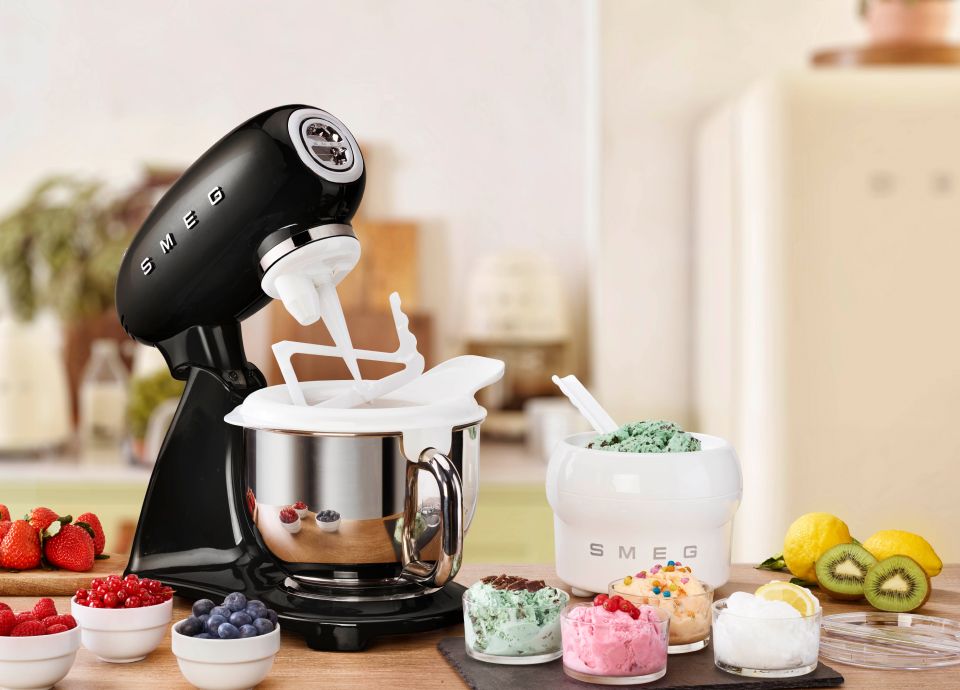 A sleek matt black food mixer. On the base sits a polished stainless steel bowl. A white partial lid sits on top through which a white ice cream mixing blade is positioned. To the right of the mixer, is Smeg's ice cream maker accessory with fresh. delicious looking, mint choc chip ice cream showing.