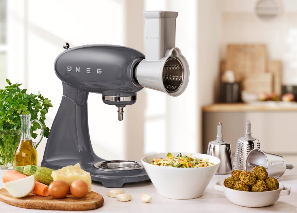 A grey smeg food mixer with a vegetable cutter accessory attached to the front, and beneath it, is a bowl of freshly cut vegetables such as carrots, peppers and cucumbers