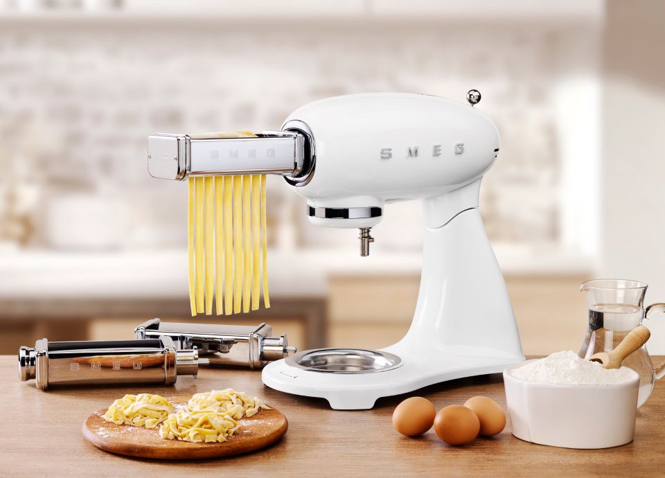 A white Smeg food mixer with an attractive Chrome pasta cutter attached to the front, and fresh pasta hanging from the attachment as it's being cut