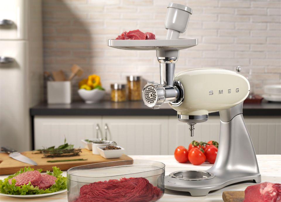 A cream retro Smeg food mixer on a kitchen worktop surrounded various meats being prepared for a BBQ. Attached to the front is a food grinder ready to prepare meat into a fine mince